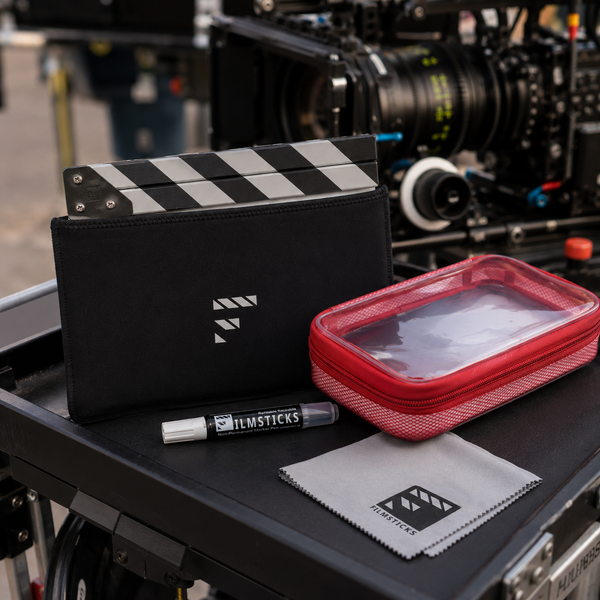 Filmsticks cleaning kit including a black clapperboard cover, red zippered case, white marker, and gray microfiber cloth, displayed on a film equipment table with a camera lens blurred in the background.